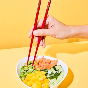 Eine weiße Bowl angerichtet mit gelben Paprika, Zucchinistücken, Edamame und Lachs. In einer Hand zwei Rote Stäbchen fassen zum Essen in die Schüssel. Der Hintergrund ist gelb.