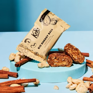 Eine Mischung aus Nüssen und Zimtstangen auf einem blauen Teller. Darauf steht eine Packung mit Bio Energy Balls, zwei davon liegend ausgepackt davor.