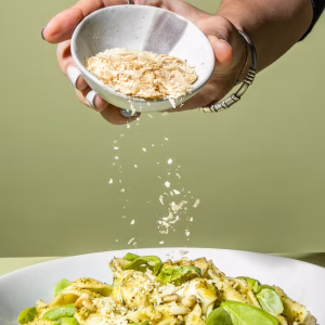 Eine Hand hält eine kleine, weiße Schale mit hellen Flocken über einen tiefen Teller mit Pasta und frischen grünen Blättern. Die Flocken werden aus der Schale auf das Gericht gestreut. Die Person trägt einen dunklen Pullover und ein Armband. Der Hintergrund ist einfarbig und hellgrün.