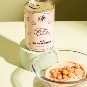 Eine Dose Bio Kichererbsen steht auf einem hellgrünen Sockel. Davor eine Schüssel mit Aufstrich und Kichererbsen