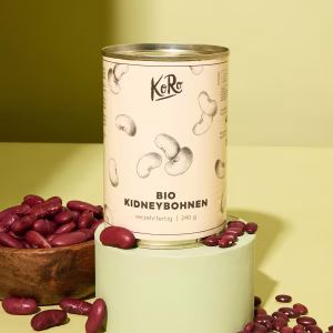 Eine Dose Bio Kidneybohnen steht auf einem hellgrünen Sockel. Davor und daneben liegen Kidneybohnen verteilt vor grünem Hintergrund,