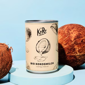 Eine Dose Kokosmilch Bio steht auf einem blauen Sockel. Eine Kokosmilch liegt daneben vor blauem Hintergrund