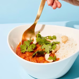 Eine weiße Bowl ist angerichtet mit Reis, Fleisch in Soße und frischer Petersilie. Vor blauem Hintergrund.