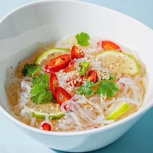 Eine weiße Bowl ist angerichtet mit Konjaknudeln und gemischtem Gemüse. Dahinter ein blauer Hintergrund