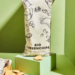 Eine Packung Bio Linsenchips mit Meersalz steht auf einem grünen Sockel. Davor liegen einzelne Linenschips verteilt vor grünem Hintergrund