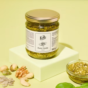Ein Glas Pesto Verde vegan steht auf einem grünen Absatz. Der Deckel ist golden. Davor steht eine Schüssel mit Pesto und frischen Basilikum, außerdem Nüsse dekoriert vor grünem Hintergrund