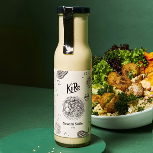 Eine Flasche Sesam Soße befindet sich neben einem Teller mit verschiedenen Speisen vor grünem Hintergrund.