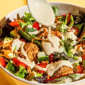 Eine helle Schüssel steht auf einem gelben Untergrund und ist gefüllt mit einem Salat. Zu sehen sind Tofu Rosso, Karotten, grüner Salat, rote Paprika, Gruken und ein weißes Dressing. Über der Schüssel eine Löffel mit Dressing. Das Bild ist klar und farbenfroh, die Zutaten sind gut erkennbar. Der Hintergrund ist einfarbig blau.