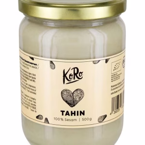 Ein durchsichtiges Glas mit goldenem Deckel beschriftet mit Tahin. Der Hintergrund ist weiß.