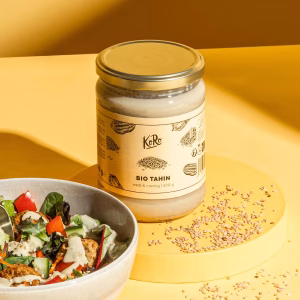 Ein Glas Bio Tahini steht aufrecht auf einem gelben Podest vor einem gelben Hintergrund. Das Tahini ist in einem Glas mit goldenen Deckel zu sehen. Auf dem Podest und rund um das Glas liegt verstreut Sesamkörner. Links daneben steht eine weiße Schüssel mit einem bunten Salat angerichtet mit einem weißen Dressing. Das Produktetikett enthält Informationen zu Zutaten und Nährwerten. Die Bildgestaltung ist hell und modern, die Farben sind kräftig und klar.