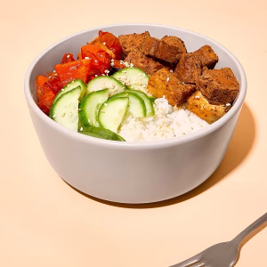 Eine weiße Schüssel ist angerichtet mit einer Bowl aus Gurken, roten Paprika, Reis und Tofu Rosso, dekoriert mit Sesam. Davor liegt eine Gabel. Der Hintergrund ist leicht rosa.