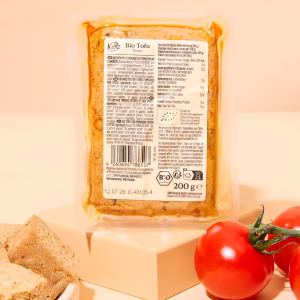 Eine Packung Bio Tofu Rosso steht aufrecht auf einem rosafarbenen Podest vor einem rosaHintergrund. Der Tofu ist in einer durchsichtigen Kunststoffverpackung zu sehen. Auf dem Podest und rund um die Packung liegen lose Tomate und Tofu-Stücke. Das Produktetikett enthält Informationen zu Zutaten und Nährwerten. Die Bildgestaltung ist hell und modern, die Farben sind kräftig und klar.
