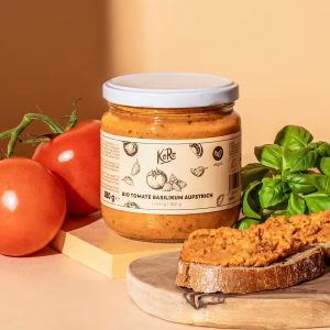 Eine durchsichtiges Glas mit weißem Deckel mit Bio Tomate Basilikum Aufstrich. Das Glas steht aufrecht vor einem orangenfarbenen Hintergrund. Das Etikett zeigt die Produktbezeichnung, Zutaten und Nährwerte. Vor der Packung liegen einzelne Brot-Stücke mit dem Aufstrich. Links im Bild liegen zwei Tomaten, rechts frische Basilikumzweige. Das Gesamtbild wirkt hell und modern, die Produkte werden klar erkennbar präsentiert.