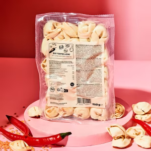 Eine Packung Bio Tortelloni mit Linsen und Chili steht aufrecht auf einem rosafarbenen Podest vor einem roten Hintergrund. Die Tortelloni sind in einer durchsichtigen Kunststoffverpackung zu sehen. Auf dem Podest und rund um die Packung liegen lose Tortelloni, einige rote Chilischoten und verstreute gelbe Linsen. Das Produktetikett enthält Informationen zu Zutaten und Nährwerten. Die Bildgestaltung ist hell und modern, die Farben sind kräftig und klar.