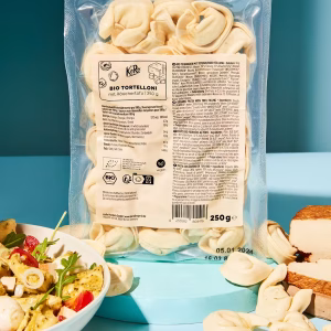 Eine transparente Plastikverpackung mit Bio Tortelloni, gefüllt mit Räuchertofu. Die Verpackung ist 250 Gramm schwer und steht aufrecht vor einem hellblauen Hintergrund. Das Etikett zeigt die Produktbezeichnung, Zutaten und Nährwerte. Vor der Packung liegen einzelne Tortelloni-Stücke verstreut. Links im Bild steht eine Schüssel mit einem frischen Salat aus Tortelloni, Tomaten, Rucola und weißen Würfeln (vermutlich Tofu). Rechts unten ist ein Block Räuchertofu und weiteres Gemüse zu sehen. Das Gesamtbild wirkt hell und modern, die Produkte werden klar erkennbar präsentiert.